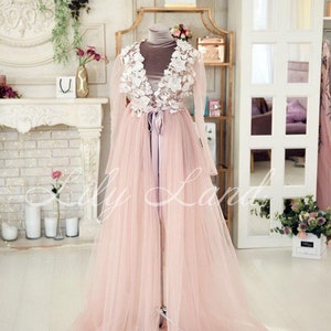 May include: A long, pink tulle gown with a white lace bodice and a satin sash. The gown is on a mannequin in a room with a fireplace and a mirror.
