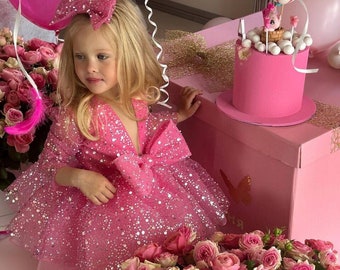 May include: A young girl in a pink sparkly dress with a large pink bow on her head stands in front of a pink cake with a hot air balloon topper. The cake is on a pink box with a butterfly design. There are pink roses and feathers in the foreground.