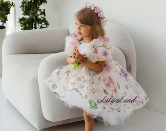 Op de afbeelding: Een jong meisje draagt een witte jurk met een bloemenprint en een bloemenkroon. De jurk heeft een tule rok en pofmouwen. Het meisje staat op een witte fauteuil en kijkt naar de zijkant. De tekst "Lily Land" is zichtbaar op de jurk.