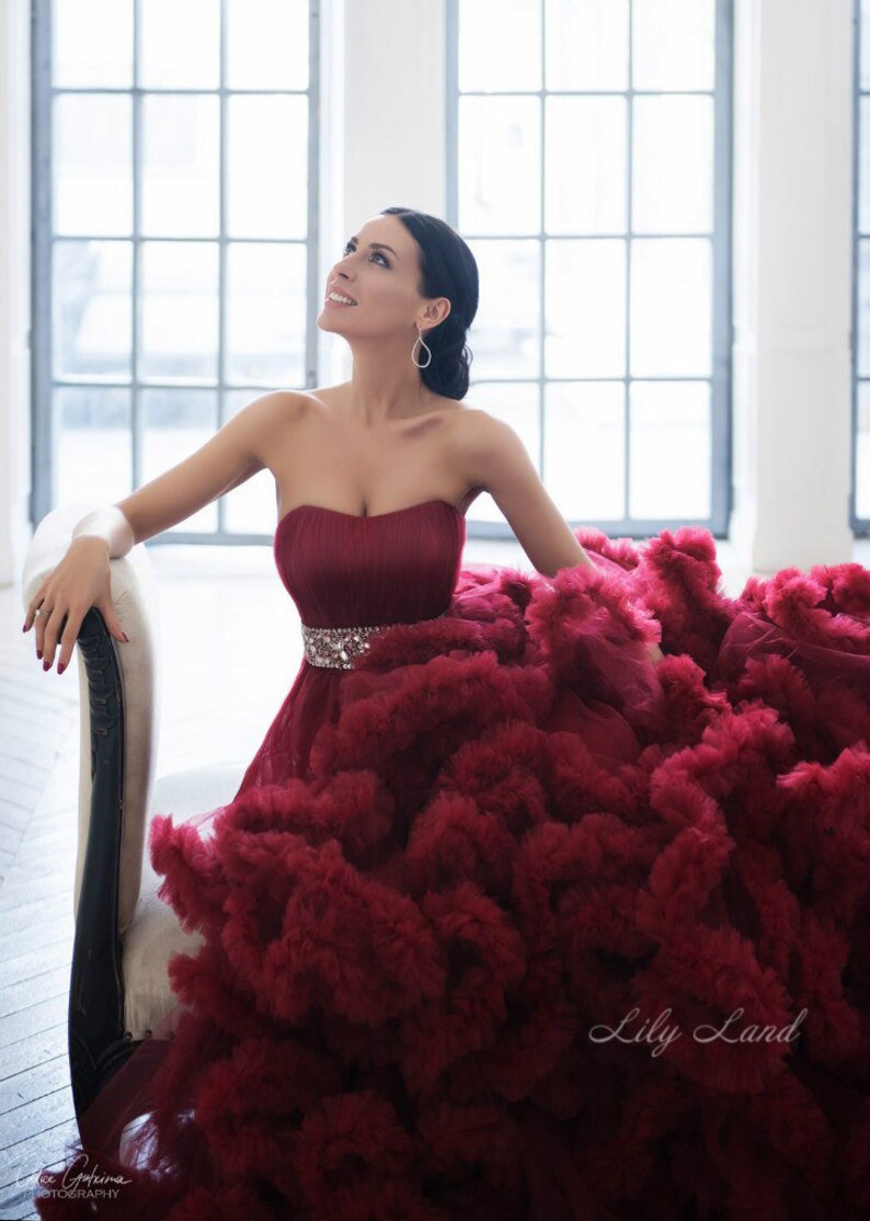 Op de afbeelding: Een vrouw in een strapless, bordeauxrode jurk met een geruite rok en een glinsterende riem. De jurk is erg vol en de vrouw zit op een witte stoel. De tekst "Lily Land" is zichtbaar in de rechterbenedenhoek van de afbeelding.