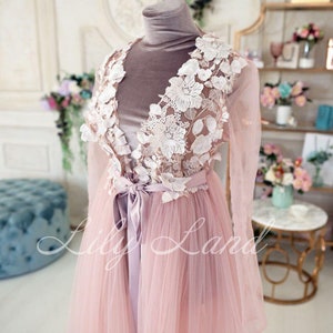 May include: A long, pink tulle gown with a white lace overlay and a satin sash. The gown has long sleeves and a V-neckline. The lace overlay features delicate floral designs.