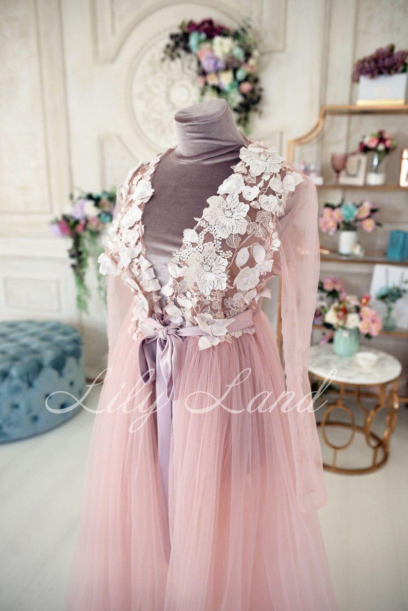 May include: A long, pink tulle gown with a white lace overlay and a satin sash. The gown has long sleeves and a V-neckline. The lace overlay features delicate floral designs.