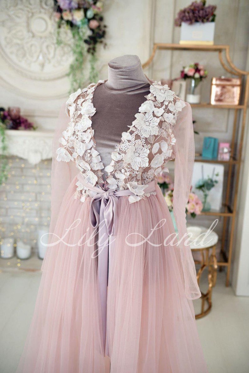 May include: A long, pink tulle gown with a white lace overlay and a satin sash. The lace features delicate floral designs. The gown is on a mannequin.