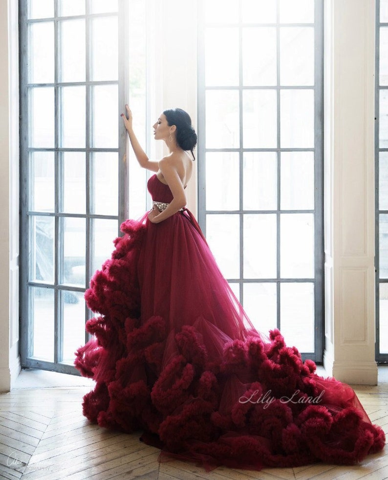 Op de afbeelding: Een vrouw die een strapless, bordeauxrode tulejurk draagt met een gerimpelde rok en een glinsterende riem. De jurk heeft een lange sleep en is erg vol. De vrouw staat voor een raam met een witte achtergrond.