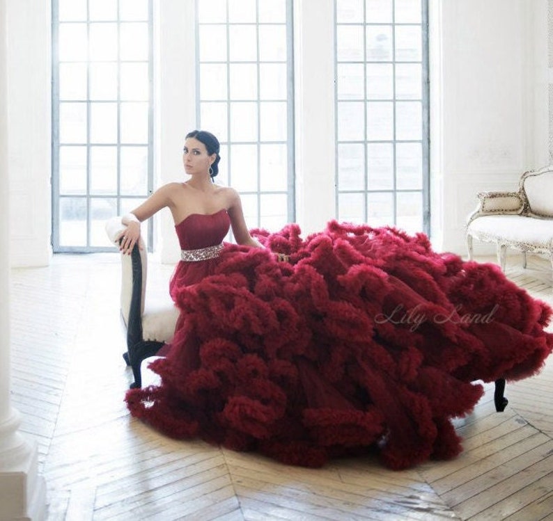 Op de afbeelding: Een vrouw in een strapless, bordeauxrode tulejurk met een gerimpelde rok. De jurk heeft een kralenband en de vrouw zit op een witte stoel. De achtergrond is een witte kamer met grote ramen.