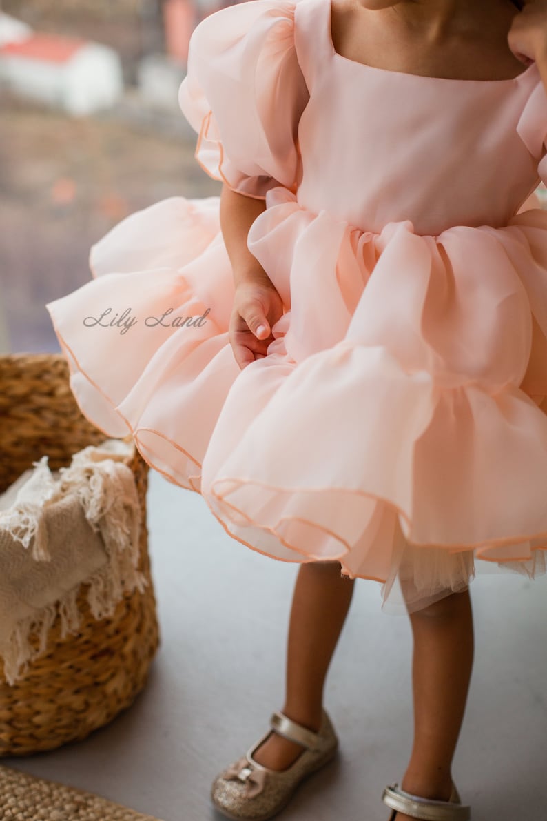 May include: A young girl wearing a peach-colored tulle dress with a ruffled skirt and puffed sleeves. The dress has a square neckline and is decorated with a delicate trim. The girl is standing on a light-colored floor.