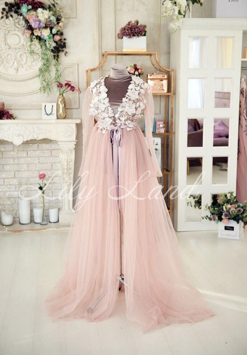 May include: A long, pink tulle gown with a white lace bodice and a satin sash. The gown is on a mannequin in a room with a fireplace and a mirror.