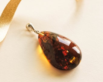 May include: A teardrop-shaped amber pendant with a silver bail. The amber is a deep brown color with inclusions of black and gold.