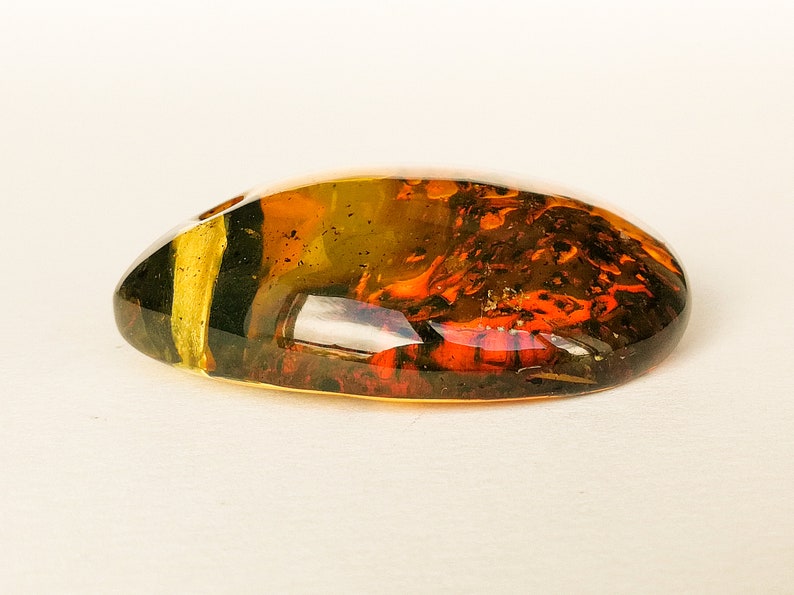 May include: A large, oval-shaped piece of amber with a mix of brown, orange, and green colors. The amber has a translucent quality, and you can see through it to some extent.