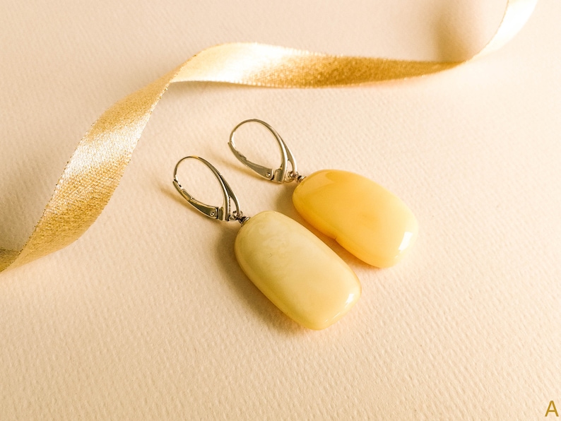 Może przedstawiać: Para srebrnych kolczyk&oacute;w z ż&oacute;łtymi kamieniami bursztynowymi. Kamienie mają owalny kształt i gładkie, polerowane wykończenie.
