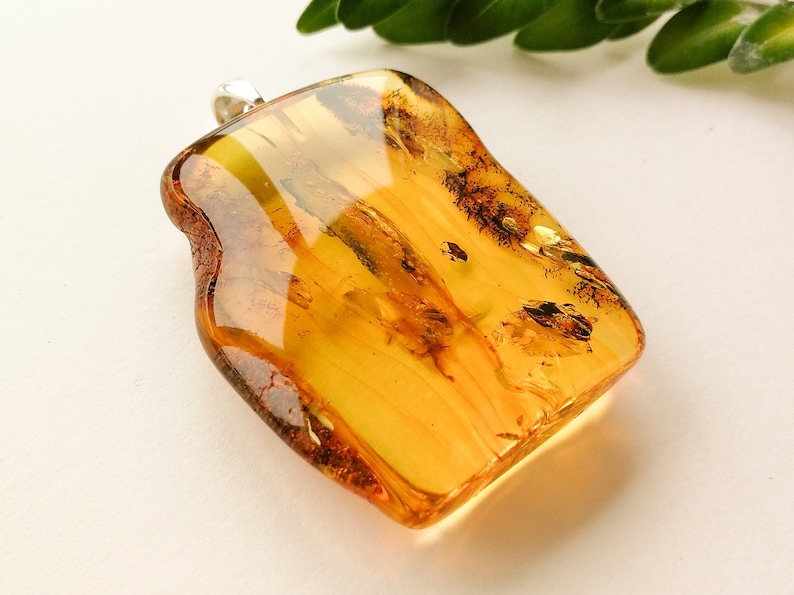 May include: A large, rectangular amber pendant with a silver bail. The amber is a warm, honey color with streaks of brown and gold. The pendant is smooth and polished.