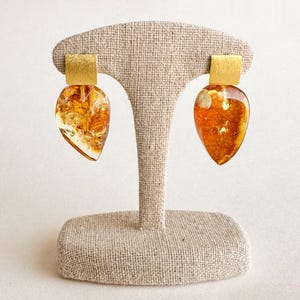 May include: A pair of amber earrings with gold accents. The earrings are teardrop shaped and have a natural, organic look. They are displayed on a beige fabric stand.