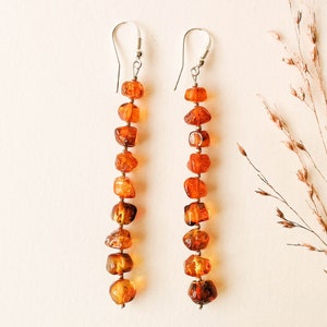 May include: A pair of long amber earrings with silver hooks. Each earring features a string of amber beads in a warm, honey-like color.