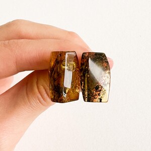 May include: Two rectangular amber-coloured stones with a polished, glossy finish. The stones have a mix of golden and brown hues with some areas of copper-coloured inclusions. The stones are held in a hand.