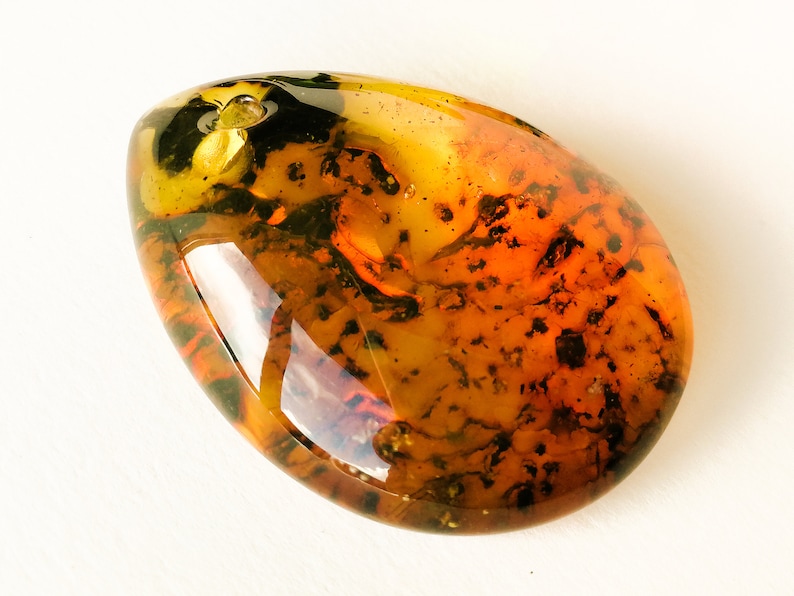May include: A teardrop-shaped amber pendant with a dark brown and green inclusion. The amber is translucent and has a warm, golden hue.