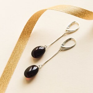 May include: A pair of silver dangle earrings with dark oval-shaped gemstones. The earrings are hanging from a gold ribbon.