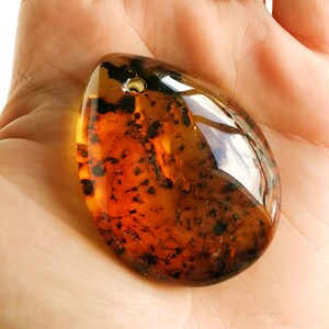 May include: A teardrop-shaped amber pendant with a hole drilled through the top. The amber is a deep orange color with dark brown and black inclusions.