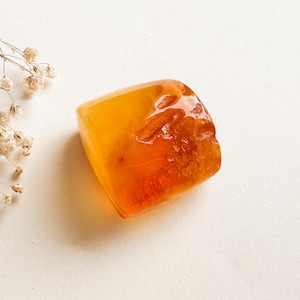 May include: A polished, translucent amber stone with a warm orange and brown color gradient. The amber has a rounded, rectangular shape and a slightly rough texture. Dried, delicate white flowers are in the background.