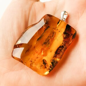 May include: A large, amber-colored pendant with a silver bail. The amber is translucent and has a warm, golden hue. The pendant is irregularly shaped and has a smooth, polished surface.