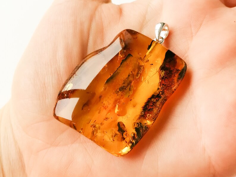 May include: A large, amber-colored pendant with a silver bail. The amber is translucent and has a warm, golden hue. The pendant is irregularly shaped and has a smooth, polished surface.
