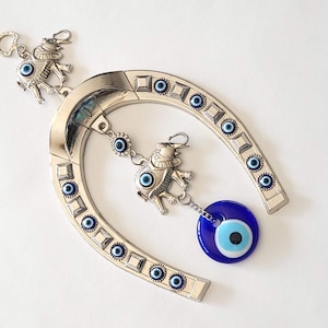 Op de afbeelding: Een zilveren hoefijzer met blauwe boze oogcharms. Het hoefijzer heeft een rij blauwe boze ogen langs de rand. Er zijn twee olifantcharms met blauwe boze ogen die aan het hoefijzer hangen. Een van de olifantcharms heeft een groot blauw boze oog dat eraan hangt.