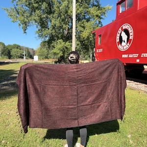 May include: A person holding up a large, rectangular, maroon-colored blanket with a fringed edge. The blanket is displayed outdoors on a sunny day, with a red train car in the background.