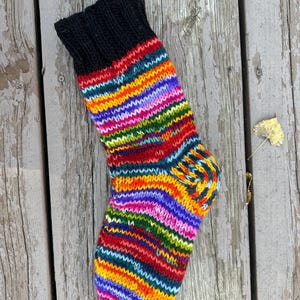 May include: A hand-knit sock featuring a solid black ribbed cuff and a vibrant, multi-colored striped pattern throughout the foot and leg. The stripes consist of alternating bands of bright orange, yellow, green, purple, and red yarn.