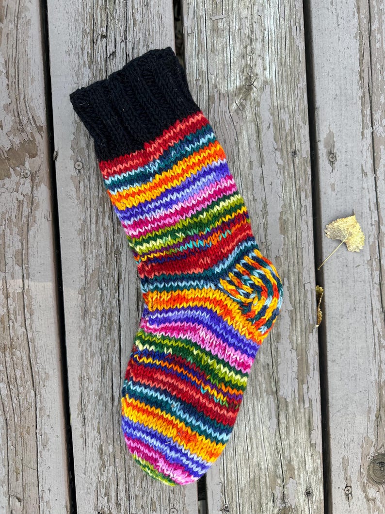 May include: A hand-knit sock featuring a solid black ribbed cuff and a vibrant, multi-colored striped pattern throughout the foot and leg. The stripes consist of alternating bands of bright orange, yellow, green, purple, and red yarn.