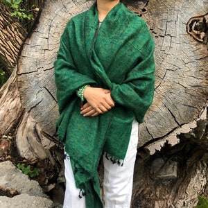 May include: A green shawl with a fringed edge. The shawl is draped over the shoulders. The person is wearing white pants and a green beaded bracelet. The background is a large tree trunk.