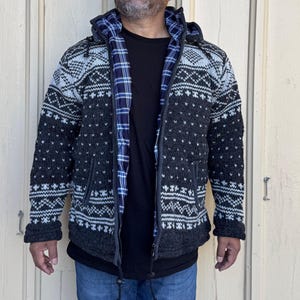 Op de afbeelding: Een hoodie met ritssluiting in grijstinten en zwart, met een wit geometrisch patroon. De binnenvoering is een blauwe geruite stof. De trui heeft een ritssluiting aan de voorkant en zakken. De persoon draagt een zwart T-shirt en een blauwe jeans.