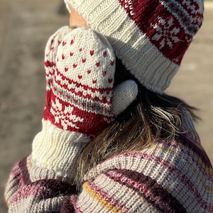 May include: Cream-colored knitted mittens and a matching hat with red and gray geometric patterns. The mittens have a heart-shaped design. The hat and mittens are part of a winter clothing set. The person is also wearing a sweater with a striped pattern.
