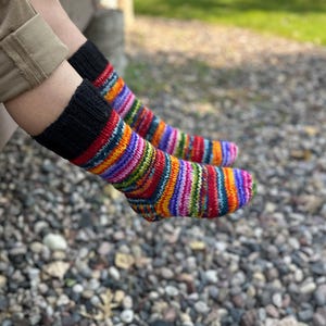 May include: A pair of hand-knitted socks featuring a vibrant, multi-colored striped pattern in shades of red, orange, yellow, green, blue, and purple. The socks have a solid black ribbed cuff at the top and a textured knit construction.