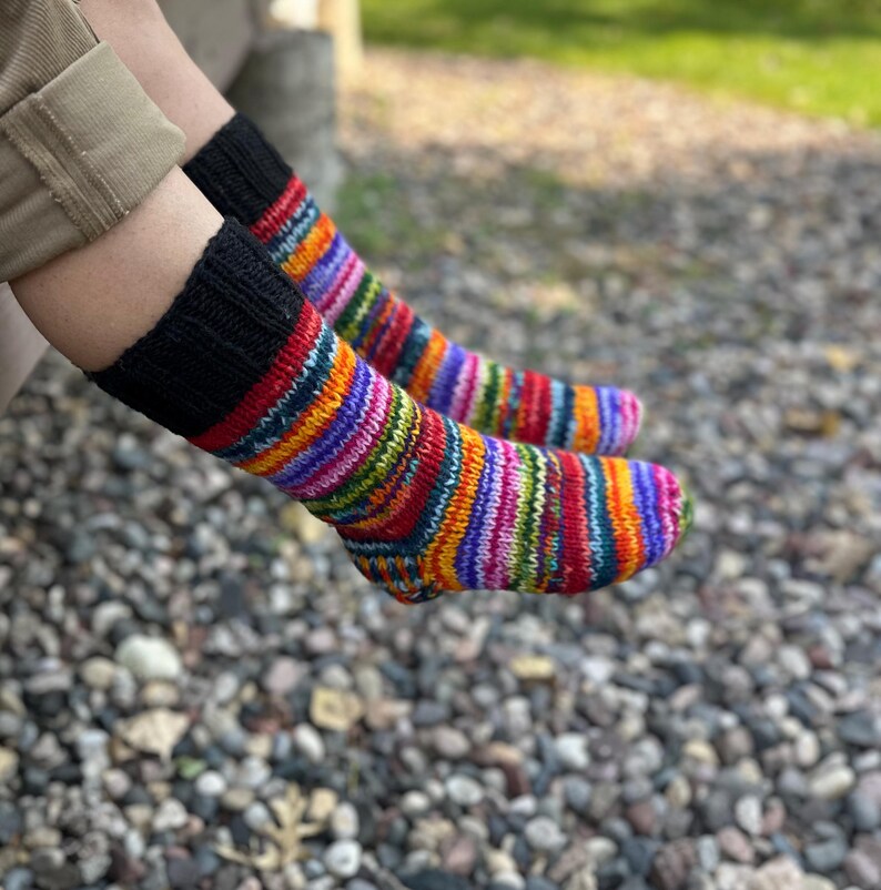 May include: A pair of hand-knitted socks featuring a vibrant, multi-colored striped pattern in shades of red, orange, yellow, green, blue, and purple. The socks have a solid black ribbed cuff at the top and a textured knit construction.