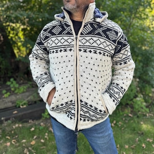 Può includere: Un maglione con zip color crema con un motivo geometrico nero. Il maglione ha un cappuccio con fodera a quadri blu e neri e due tasche anteriori. La persona indossa jeans blu.
