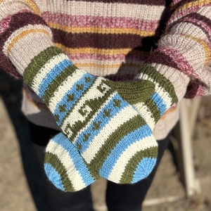 May include: A pair of knitted mittens with a striped pattern in white, blue, and green. The mittens are folded together and held in the hands of a person wearing a striped sweater.