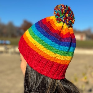 May include: A rainbow striped knit beanie with a red base and a colorful pom pom on top.