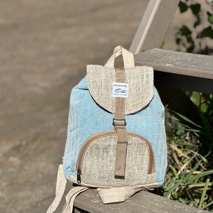 Puede incluir: Una pequeña mochila de cáñamo azul y beige con un patrón de chevron. La mochila tiene un solo bolsillo con cremallera en la parte delantera y una correa ajustable marrón. La etiqueta en la parte delantera dice "Handmade, Respect the Earth".