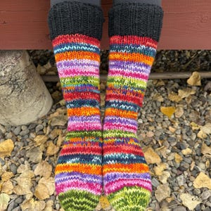 May include: A pair of hand-knitted socks featuring a vibrant, multi-colored striped pattern in shades of orange, purple, pink, green, and blue. The socks have a solid black ribbed cuff at the top and a green knit toe section.