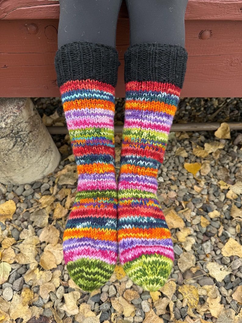 May include: A pair of hand-knitted socks featuring a vibrant, multi-colored striped pattern in shades of orange, purple, pink, green, and blue. The socks have a solid black ribbed cuff at the top and a green knit toe section.
