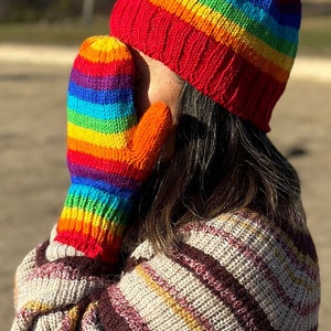 May include: A person wearing a rainbow striped knitted hat and matching mittens. The hat and mittens are red, orange, yellow, green, blue, indigo, and violet.