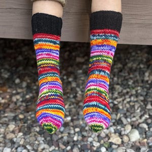 May include: A pair of hand-knitted socks featuring a vibrant, multi-colored striped pattern in shades of red, orange, yellow, green, blue, and purple. The socks have a solid black ribbed cuff at the top and a standard knit texture throughout.