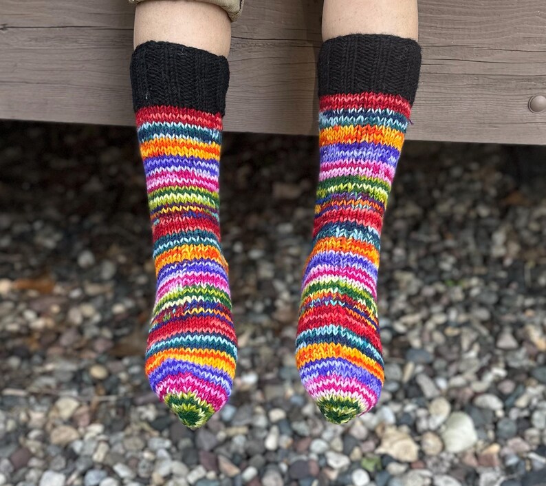 May include: A pair of hand-knitted socks featuring a vibrant, multi-colored striped pattern in shades of red, orange, yellow, green, blue, and purple. The socks have a solid black ribbed cuff at the top and a standard knit texture throughout.