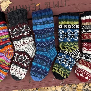 May include: Five pairs of hand-knitted wool socks arranged in a row. The socks feature various patterns including multi-colored horizontal stripes, intricate geometric designs in black, white, blue, green, and deep red, and ribbed cuffs.