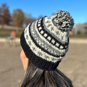 Könnte beinhalten: Eine gestrickte Beanie-Mütze mit einem geometrischen Muster in Schwarz, Weiß und Grau. Die Mütze hat einen schwarzen Rand und einen flauschigen Bommel oben. Die Mütze ist für kaltes Wetter konzipiert und eignet sich für Outdoor-Aktivitäten.