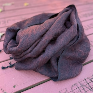 Peut inclure: Une écharpe bordeaux foncé avec un aspect tissé texturé. L'écharpe est drapée et pliée, révélant un mélange de teintes bordeaux et violet foncé. De petites glands noirs sont visibles au bord de l'écharpe. L'écharpe semble douce et chaude.