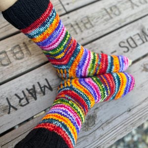 May include: A pair of hand-knitted socks featuring a vibrant, multi-colored striped pattern in shades of red, orange, yellow, green, blue, and purple. The socks have a solid black ribbed cuff at the top and a standard heel and toe construction.