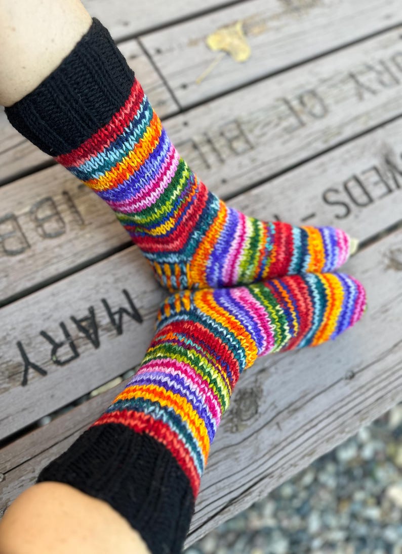 May include: A pair of hand-knitted socks featuring a vibrant, multi-colored striped pattern in shades of red, orange, yellow, green, blue, and purple. The socks have a solid black ribbed cuff at the top and a standard heel and toe construction.