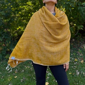 May include: Mustard yellow shawl with a textured appearance. The shawl is draped over the shoulders and has a fringed edge. The fabric appears soft and warm, suitable for cooler weather. A small tag is visible on the corner of the shawl.