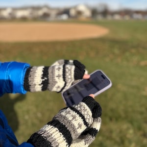 Puede incluir: Guantes sin dedos tejidos a mano en negro, blanco y gris. Los guantes presentan un diseño de estrellas y rayas. La persona sostiene un teléfono inteligente con una funda morada. El fondo es un campo de hierba y un campo de béisbol.
