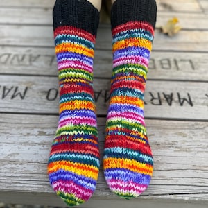 May include: A pair of hand-knitted socks featuring a vibrant, multi-colored striped pattern in shades of red, orange, yellow, green, blue, and purple. The socks have a solid black ribbed cuff at the top and a standard knit texture throughout.